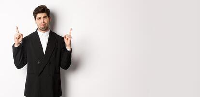 Portrait of reluctant and sad handsome man, complaining and pointing fingers up at something bad, looking jealous, standing over white background photo