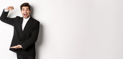 Portrait of excited handsome man in suit, shaping big object on copy space and smiling amazed, holding something, standing over white background photo