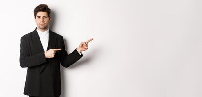 Image of handsome businessman in black suit, pointing fingers right and looking at camera, standing against white background photo