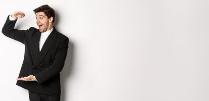 Portrait of excited handsome man in suit, shaping big object on copy space and smiling amazed, holding something, standing over white background photo