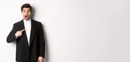 Concept of new year party, celebration and lifestyle. Portrait of surprised handsome guy in black suit, pointing at himself and looking amazed, being chosen, standing over white background photo