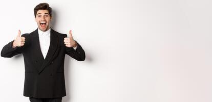 Concept of new year party, celebration and lifestyle. Portrait of amazed and pleased handsome man in black suit, showing thumbs-up, like product, approve something good, white background photo