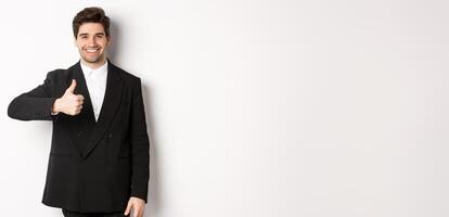 Portrait of handsome and confident male realtor, showing thumb-up and smiling, guarantee quality and recommending company, standing over white background photo