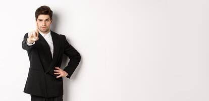 Portrait of serious handsome man in business suit, showing one finger to prohibit or decline something, telling to stop, disagree with you, standing over white background photo