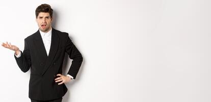 Portrait of arrogant man in black suit, looking confused and disappointed, complaining about something strange, standing over white background photo