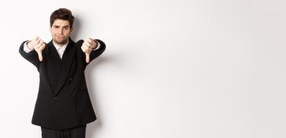 Portrait of skeptical and disappointed man in black suit, frowning upset, showing thumbs-down, dislike something bad, standing over white background photo