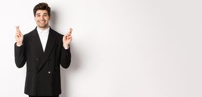 Hopeful and optimistic young businessman in suit, smiling while crossing fingers for good luck and making wish, standing over white background photo
