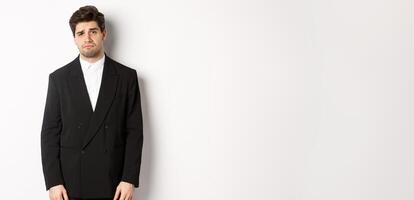 Image of gloomy and sad handsome male model in black suit, frowning and looking tired at camera, standing against white background photo