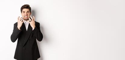 Image of angry businessman in suit, looking with furious expression and clenching fists, express hatred, standing mad over white background photo