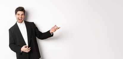 Portrait of happy man dancing at party, playing on invisible guitar and laughing, standing in black suit against white background photo