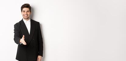 Portrait of handsome businessman in black suit, extending hand for handshake, greet business partners and smiling, welcome to company, standing over white background photo
