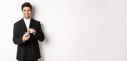 Image of handsome businessman in suit showing you app on smartphone, pointing finger at mobile screen and smiling, standing over white background photo