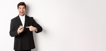 Image of cheerful businessman looking amazed, pointing at mobile phone, standing in suit over white background photo