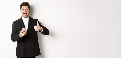 Portrait of handsome man in trendy suit showing thumbs-up in approval, using mobile phone app, smiling pleased, standing over white background photo