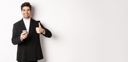 Handsome guy in trendy suit making compliment, showing thumbs-up while using mobile phone, recommending an app or online shop, standing over white background photo