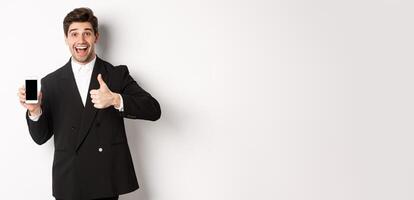 Image of handsome male entrepreneur in black suit, recommending app or online shop, showing thumbs-up and smartphone screen, standing over white background photo