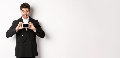Portrait of excited handsome man showing smartphone screen, looking amazed at display with advertisement, standing over white background in black suit photo