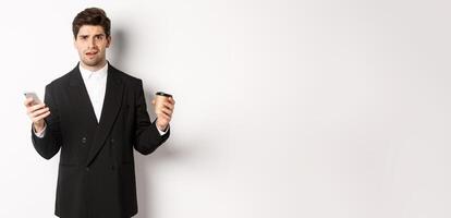 Image of confused businessman standing in a suit, holding cup of coffee and mobile phone, cant understand something, standing against white background photo