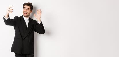 Image of handsome man in suit, having call and waving hand at smartphone camera, recording , greeting someone, standing against white background photo