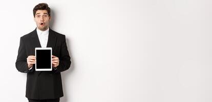 Image of surprised handsome man in black suit, showing digital tablet screen and looking amazed, standing against white background photo