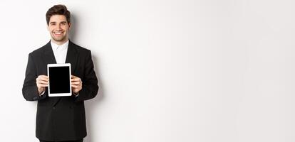 Portrait of handsome businessman in trendy suit, showing digital tablet screen and smiling, standing against white background photo