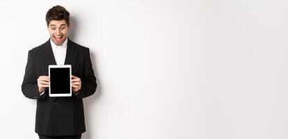 Portrait of amazed handsome man in black suit, showing digital tablet screen with excitement, standing over white background photo