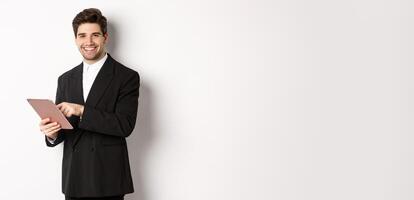 Image of handsome businessman in black suit, using digital tablet and smiling, standing against white background photo
