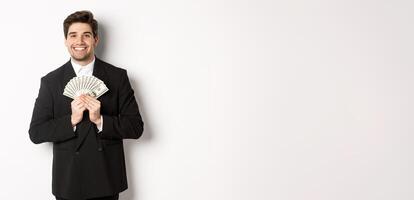 Portrait of happy and pleased handsome man in suit, hugging money and looking satisfied, standing over white background photo