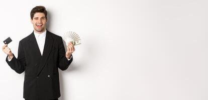 Image of confident businessman in black suit, smiling pleased and winking, holding money and credit card, standing against white background photo