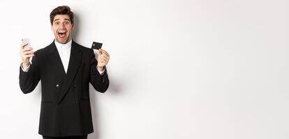 Handsome businessman in black suit smiling, showing credit card and money, looking amazed, standing against white background photo