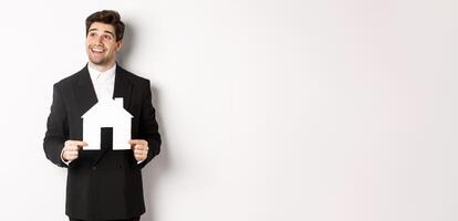 Image of handsome investor in black suit, holding house maket and looking at upper left corner thoughtful, standing against white background photo