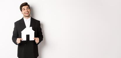 Portrait of handsome man in suit searching for home, holding paper house and looking at upper right corner dreamy, standing over white background photo