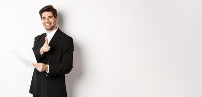 Image of handsome businessman in black suit, holding document and pointing finger at camera, praising good job, standing against white background photo