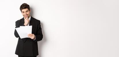 Image of handsome businessman in black suit, looking pleased at documents, reading report and smiling, standing against white background photo