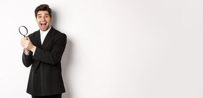 Handsome businessman in black suit, holding magnifying glass and smiling, found something, standing against white background photo