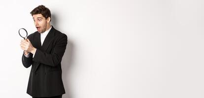 Handsome businessman in black suit, holding magnifying glass and smiling, found something, standing against white background photo
