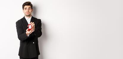Portrait of handsome man making a proposal, asking to marry him, showing ring, standing in black suit against white background photo