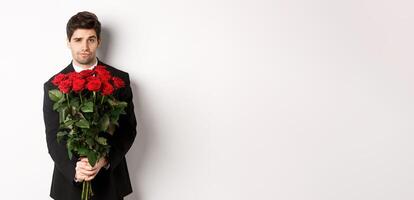Image of handsome boyfriend in black suit, holding bouquet of red roses and smiling, being on a date, standing over white background photo