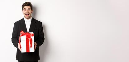 Concept of christmas holidays, celebration and lifestyle. Image of handsome guy in black suit looking excited, have a gift, standing against white background photo