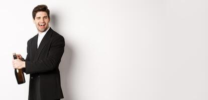 Portrait of attractive man in black suit, winking at camera and opening bottle of champagne, celebrating new year, standing against white background photo