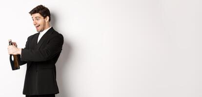 Portrait of attractive man in black suit, winking at camera and opening bottle of champagne, celebrating new year, standing against white background photo