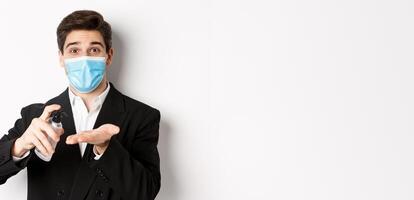 Concept of covid-19, business and social distancing. Image of handsome businessman in trendy suit and medical mask, cleaning hands with hand sanitizer, standing over white background photo