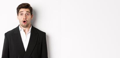 Close-up of attractive man in black suit, looking surprised and impressed at advertisement, standing over white background photo