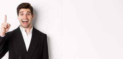 Close-up of handsome businessman in black suit, smiling amazed, showing number one, standing over white background photo