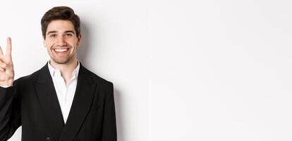 Close-up of handsome businessman in black suit, smiling amazed, showing number two, standing over white background photo