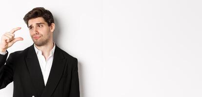 Close-up of handsome businessman in trendy suit, showing something small with disappointment, standing against white background photo