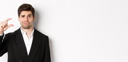Close-up of handsome businessman in trendy suit, showing something small and looking disappointed, standing against white background photo