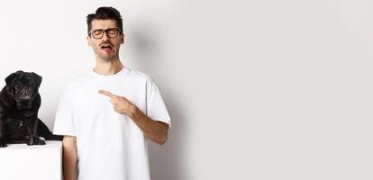 Upset crying man pointing at cute black pug and sobbing, complaining on his pet, standing sad against white background photo