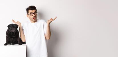 Portrait of confused hipster guy dog owner shrugging, standing near cute black pug pet, white background photo