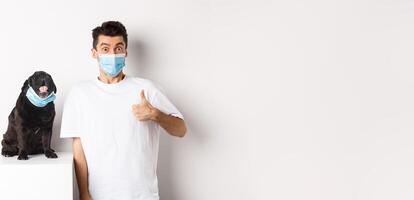 Covid-19, animals and quarantine concept. Image of funny young man and small dog in medical masks, owner showing thumb up in approval, praise something, white background photo